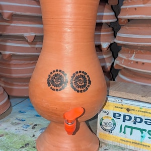 Puede incluir: Un dispensador de agua de terracota con tapa y grifo. El dispensador tiene dos diseños florales negros y está rodeado de otras ollas de terracota. Probablemente se utiliza para almacenar y dispensar agua.