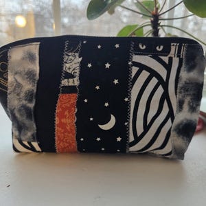 May include: A black and white patchwork cosmetic bag with a zipper closure. The bag features various fabric patterns, including a skull, stars, a crescent moon, and striped and patterned sections. The bag is rectangular and sits on a white surface.