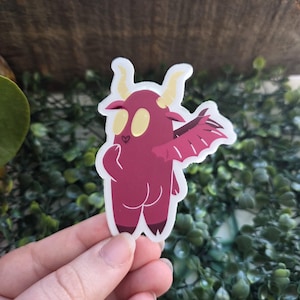 May include: A sticker featuring a cartoon demon with a maroon body, yellow eyes, and cream-colored horns. The demon has a heart-shaped nose and a single wing. The sticker is held by a hand.
