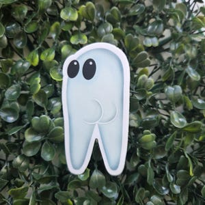 May include: A light blue cartoon tooth sticker with black eyes and a smiling mouth. The sticker has a white border and is set against a backdrop of green leaves.