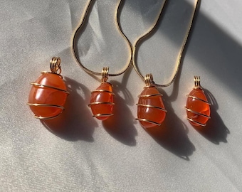 May include: Four gold-colored chain necklaces with orange gemstone pendants. Each pendant is an oval-shaped orange stone wrapped in gold-colored wire. The necklaces are displayed on a white, textured surface.