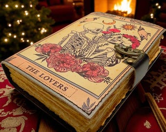 Op de afbeelding: Een tarotkaartboek met een zwarte leren riemsluiting. De kaft toont een skeletpaar, bloemen en de woorden "THE LOVERS". Het boek heeft verouderde, vergeelde pagina's en ligt op een rood oppervlak met patronen.