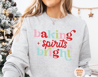 May include: A light gray sweatshirt with the text "baking + spirits bright" in colorful letters. The text is in pink, red, green, and blue. The sweatshirt is a crew neck style.
