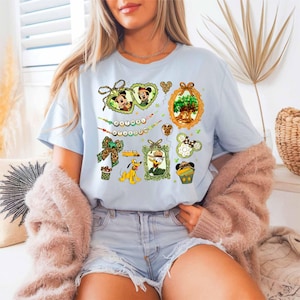 A woman wearing a light blue t-shirt with various Disney-themed illustrations, including Mickey Mouse, Minnie Mouse, and other characters, along with a floral design. She is also wearing a pink cardigan and denim shorts.