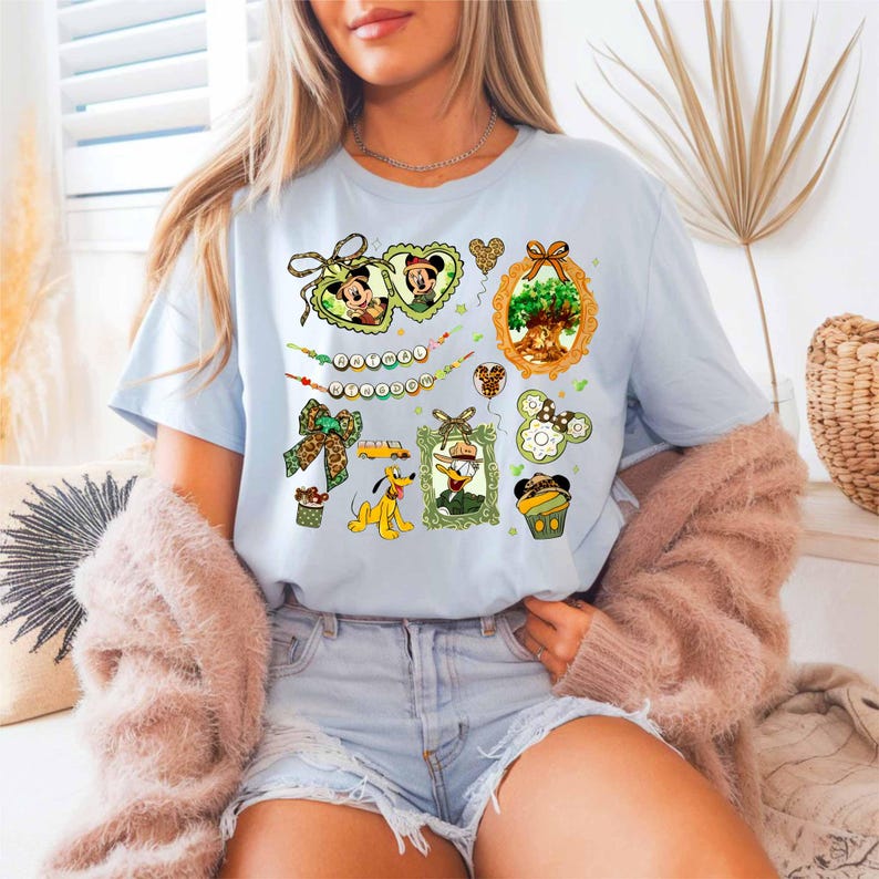 A woman wearing a light blue t-shirt with various Disney-themed illustrations, including Mickey Mouse, Minnie Mouse, and other characters, along with a floral design. She is also wearing a pink cardigan and denim shorts.