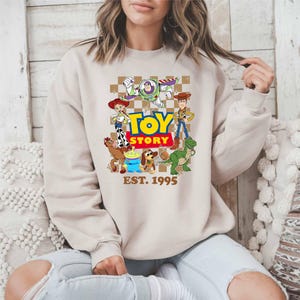 A woman wearing a beige sweatshirt with a graphic of characters from the Toy Story movie.