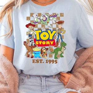 A woman wearing a light blue t-shirt with a graphic of characters from the Toy Story movie, including Woody, Buzz Lightyear, and other beloved characters. She is also wearing a pink cardigan and denim shorts.