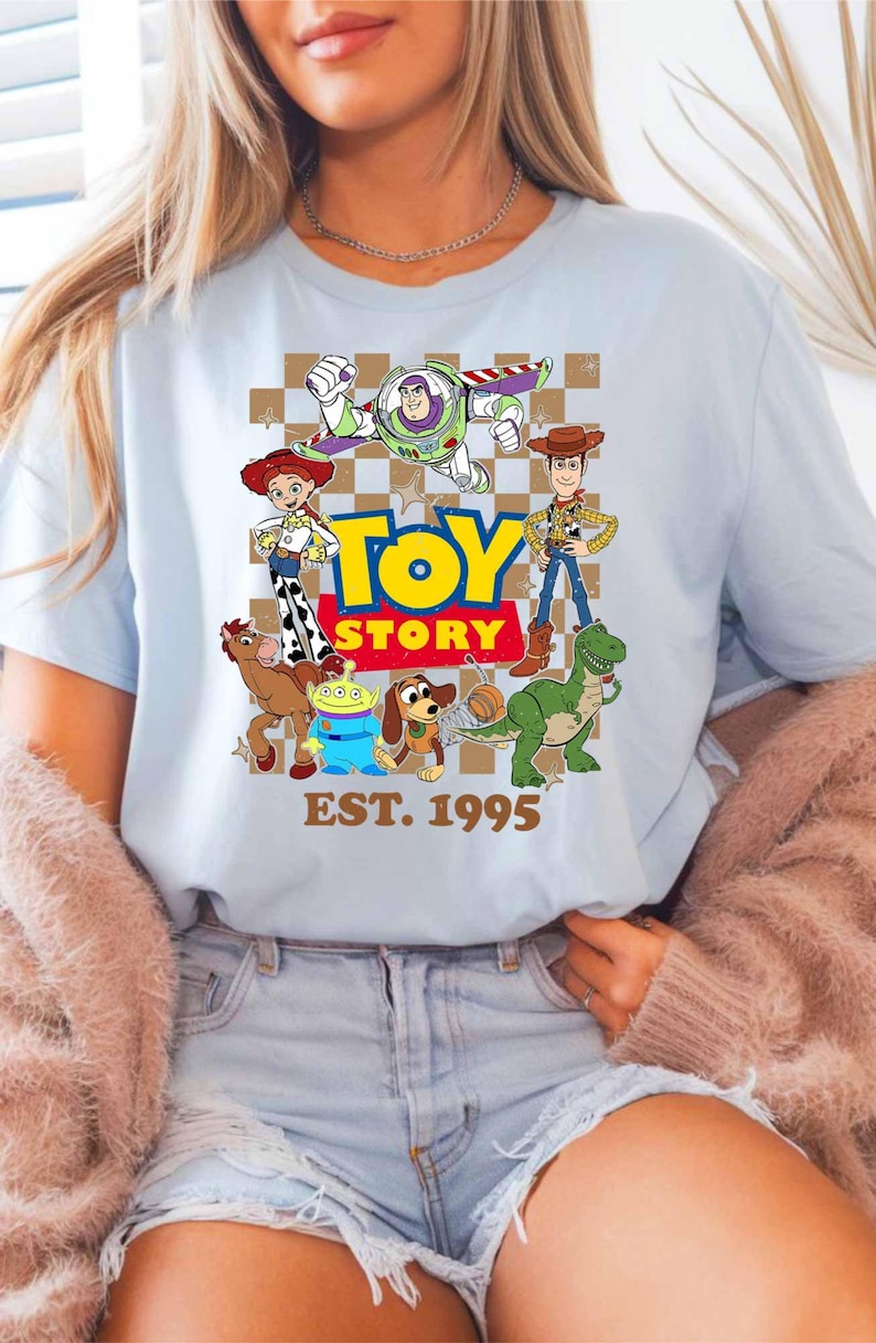 A woman wearing a light blue t-shirt with a graphic of characters from the Toy Story movie, including Woody, Buzz Lightyear, and other beloved characters. She is also wearing a pink cardigan and denim shorts.