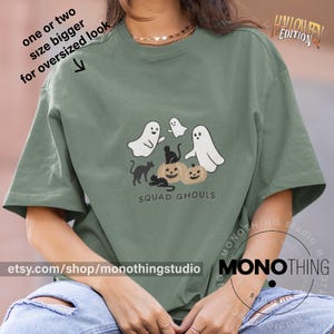 Peut inclure: T-shirt vert sauge avec un motif sur le thème d'Halloween. Le motif présente des fantômes blancs, des chats noirs et des citrouilles, avec le texte "SQUAD GHOULS". Le t-shirt porte également le texte "HALLOWEEN EDITION" et "etsy.com/shop/monothingstudio".
