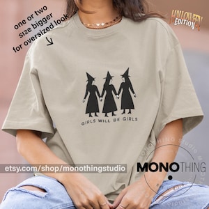 Pode incluir: Camiseta bege com um gráfico preto de três bruxas de mãos dadas, com o texto "GIRLS WILL BE GIRLS". A camisa também tem o texto "HALLOWEEN EDITION" e "MONOTHING" impressos. O URL da loja Etsy também é visível.