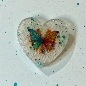 May include: A heart-shaped resin pendant with a colorful butterfly design. The butterfly has green, blue, and orange wings. The pendant is clear with glitter accents. The background is a light blue speckled surface.