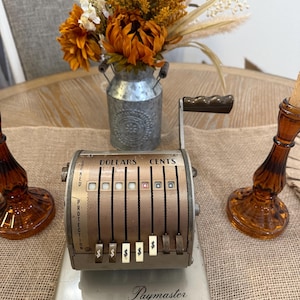 May include: An antique Paymaster cash register with a silver base and a copper-colored face. The register displays "Dollars" and "Cents" above the keys. The text "Locked Protection" is visible on the left side. The register is on a burlap surface.