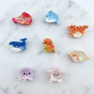 May include: A collection of eight small hair claw clips shaped like sea creatures, including a red lobster, blue dolphin, orange crab, blue whale, orange seahorse, white seashell with a pearl, purple octopus, and a pink starfish with white spots.