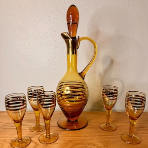 May include: A vintage amber glass decanter set with a decanter and six cordial glasses. The decanter has a gold-colored handle and stopper. Each piece is adorned with horizontal gold stripes. The set is displayed on a wooden surface.
