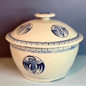 May include: Cream-colored ceramic serving bowl with a matching lid. The bowl and lid are decorated with blue bird designs and a blue geometric border. The lid has a small knob for easy handling. A decorative kitchenware item.
