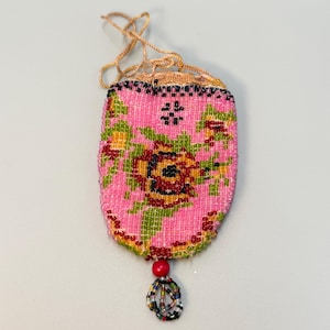 May include: A small, rectangular beaded pouch featuring a pink background with a floral pattern in green, yellow, red, and black beads. The pouch has a drawstring closure at the top and a single red bead with a multi-colored beaded tassel at the base.