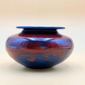May include: A handcrafted ceramic vase with a rounded body and a flared rim. The vase features a glossy finish with a blend of deep red and blue hues. The rim is a solid, dark blue. A decorative piece for the home.