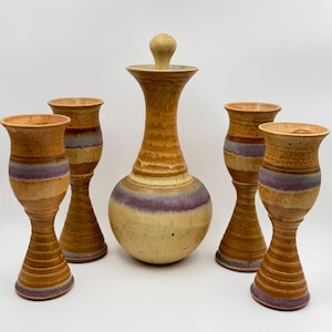 May include: A ceramic decanter with a matching stopper and four tall goblets. Each piece features a speckled tan and cream glaze with a horizontal band of muted purple around the center and base, showcasing a hand-thrown pottery construction.