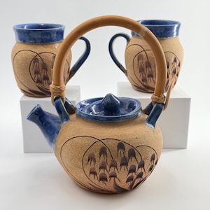 May include: A ceramic teapot and two matching mugs featuring a tan, textured clay body with blue glazed rims and lids. The pieces are decorated with dark brown painted floral motifs and the teapot includes a curved, light brown bamboo handle.