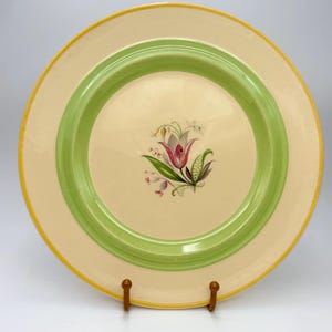May include: A round ceramic plate featuring a cream-colored center and rim, accented by a wide sage green circular band and a thin yellow outer edge. The center of the plate displays a detailed botanical illustration of a pink and white tulip.