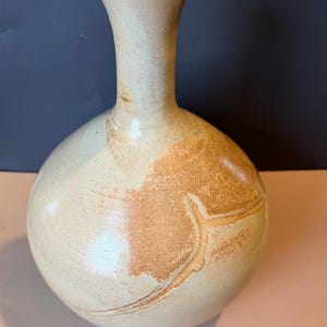 May include: A handcrafted ceramic vase with a bulbous body and a slender neck. The vase features a light beige color with brown and tan accents. The design includes a textured surface and a unique shape, suitable for home decor.