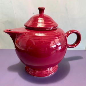 May include: A maroon ceramic teapot with a matching lid. The teapot has a rounded body, a curved handle, and a spout. The lid has a small knob on top. The teapot sits on a circular base.