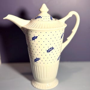 May include: Cream-colored ceramic coffee pot with a lid, spout, and handle. The pot is decorated with blue polka dots and leaf-like designs. The base has a ribbed design, and the pot stands approximately 10 inches tall.