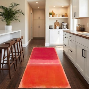 Pode incluir: Um tapete retangular com um gradiente de tons vermelhos, do vermelho escuro no topo a um laranja vibrante na parte inferior. O tapete está colocado sobre um chão de madeira escura numa cozinha, com armários brancos e uma bancada com bancos de madeira.