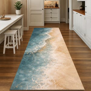 May include: A long, rectangular rug with a beach-themed design. The rug features a gradient of colors, transitioning from a deep blue representing the ocean to a sandy beige, mimicking a shoreline. White foam-like patterns add detail.