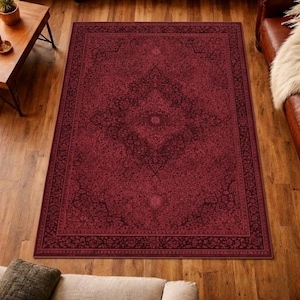 Puede incluir: Una alfombra rectangular de color burdeos con un intrincado diseño floral. La alfombra presenta un borde oscuro y un diseño central de medallón. La alfombra está colocada sobre un suelo de madera, creando un ambiente cálido.