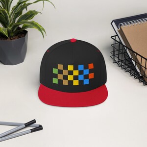 May include: A black flat-brimmed hat with a red brim and a red button on top. The hat features an embroidered design of colorful squares in green, brown, yellow, blue, and orange.