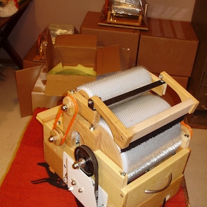 May include: A wooden wool carder with a silver metal drum and a black rubber belt. The carder is sitting on a red rug.