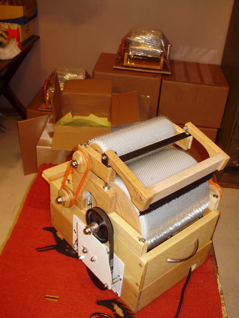 May include: A wooden wool carder with a silver metal drum and a black rubber belt. The carder is sitting on a red rug.
