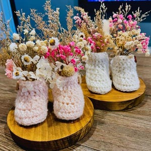 Puede incluir: Exhibición de flores secas en jarrones de ganchillo hechos a mano. Los jarrones, en tonos rosa y blanco, se asientan sobre pequeñas bases redondas de madera. Las flores, rosas, blancas y beige, crean una estética rústica y encantadora.