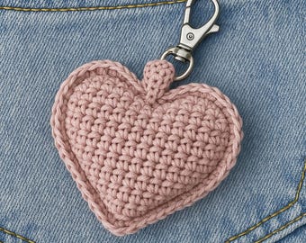 Peut inclure: Un porte-clés en forme de cœur rose crocheté avec un fermoir argenté. Le cœur est fait d'une matière tissée texturée et possède une boucle en haut pour le fermoir. Le fond est un tissu en jean.