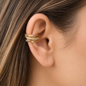 May include: A gold-toned ear cuff featuring a multi-layered, wrap-around design. The piece consists of several thin, intertwined bands, with one band encrusted with small, clear, sparkling stones and others accented by individual round stones.