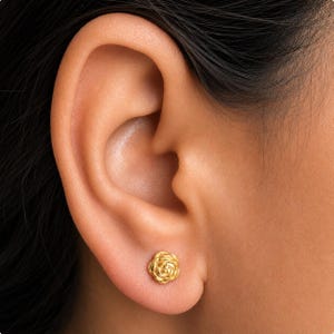 May include: A close-up of an ear with a small, gold rose stud earring. The earring is a detailed miniature of a rose, with delicate petals. The skin around the ear is smooth and light-toned, with dark hair visible.