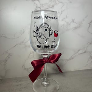 May include: A clear wine glass featuring a cartoon fish design and the text "I'M NEVER DRINKING AGAIN OH LOOK WINE". A burgundy ribbon is tied around the stem. The glass is set against a white and grey marble backdrop.