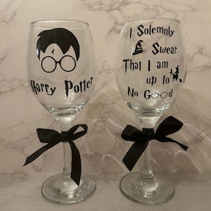May include: Two clear wine glasses with black text and graphics. One glass features a silhouette of Harry Potter and the words "Harry Potter." The other glass has the text "I Solemnly Swear That I am up to No Good." Each glass has a black bow.