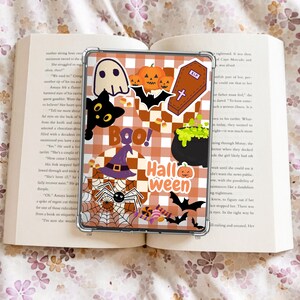 以下が含まれることがあります： ハロウィーンをテーマにしたデザインの透明な携帯電話ケース。ケースはオレンジと白のチェック柄で、お化け、黒猫、カボチャ、コウモリ、魔女の帽子、クモ、大釜、そして「Boo！」と「ハロウィーン」のイラストが描かれています。