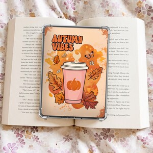 Op de afbeelding: Een transparante telefoonhoes met een herfstthema. Het ontwerp toont een roze koffiebeker met een pompoen, herfstbladeren en de woorden "Autumn Vibes" in oranje. De hoes ligt op een open boek.