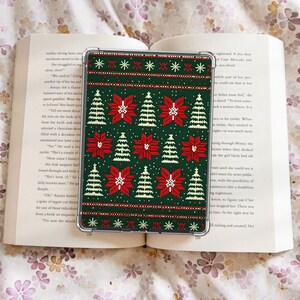 May include: A clear e-reader case with a Christmas jumper design. The pattern features red poinsettias, white snowflakes, and green Christmas trees on a dark green background. The case is on an open book.