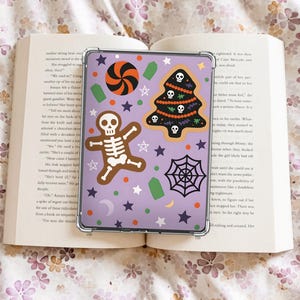 May include: A clear phone case with a Halloween-themed design, featuring a gingerbread skeleton, a spider web, a candy swirl, a Christmas tree with skulls, stars, and other festive shapes on a lavender background. The case is resting on an open book.