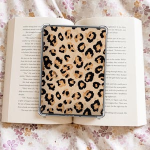 May include: A clear protective case with a leopard print design, featuring black and tan spots, rests on an open book. The book's pages are visible, and the background includes a floral pattern.