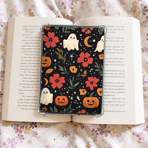 May include: A black phone case with a Halloween pattern featuring orange pumpkins, white ghosts, red flowers, and a crescent moon. The case is on an open book with floral bedding.