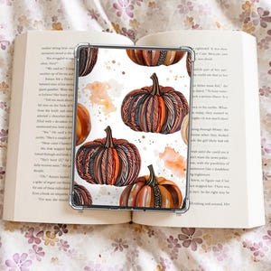 May include: A transparent phone case with a pumpkin design in orange, brown, and black watercolour. The pumpkins have detailed patterns and are set against a white background with orange splatters. The case is placed on an open book, with floral bedding visible.