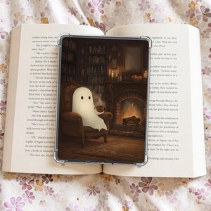 May include: A clear phone case featuring an illustration of a friendly ghost relaxing in a cozy room with a fireplace and bookshelves. The ghost is holding a cup of coffee. The case is resting on an open book with floral patterned fabric in the background.