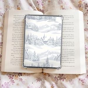 以下が含まれることがあります： 雪に覆われた家と常緑樹が描かれた冬の村の風景が特徴の透明なタブレットケース。ケースは、花柄のページがある開いた本の上に置かれています。ケースには透明な縁があります。