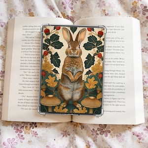 Puede incluir: Una funda transparente para teléfono con una ilustración detallada de un conejo marrón con orejas largas. El diseño incluye hojas verdes, bayas rojas y setas sobre un fondo crema. La funda se muestra sobre un libro abierto.