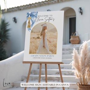 May include: A white rectangular graduation welcome sign featuring a blue watercolor bow with a gold 2025 tassel charm. The sign displays the text Welcome to Olivia Sutton's Graduation Party Lakeview High School and an arched photo frame.